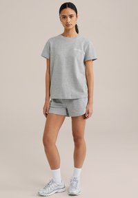 Siv osnovni pametni T-shirt in kratke hlače, z okroglim izrezom, kratkimi rokavi in majhnim natisnjenim besedilom na majici. Obuto z atletskimi supergami.