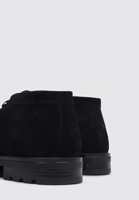 Botas de tobillo de ante negro con punta redondeada, costuras mínimas y suela de goma negra estriada. La vista trasera resalta líneas limpias.