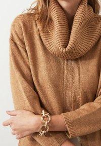 Pull oversize à col bénitier en maille marron clair avec texture côtelée. Bracelet chaîne dorée aux maillons torsadés au poignet, laissant apparaître la peau.