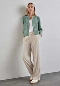Veste ajustée vert clair avec deux poches avant, associée à un pantalon large beige et des baskets blanches sur un fond gris.