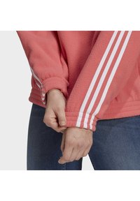 Coral fleece sweatshirt with white three-stripe accents on sleeves. Cuffs are elasticated for a snug fit, paired with dark jeans.