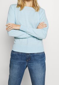 Pull bleu clair en tissu tricoté doux, présentant un col rond, des poignets et un ourlet côtelés. Associé à un jean en denim bleu foncé.