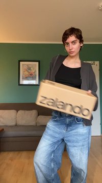 Pappeske med "zalando"-logo i fet sort skrift, holdt av en person i grå zip-up-jakke og lyseblå baggy jeans. Grønn vegg i bakgrunnen.
