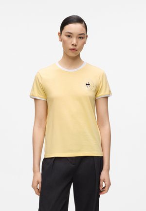 Mlada žena stoji okrenuta prema naprijed, noseći svijetložutu majicu kratkih rukava s bijelim obrubom i malim grafičkim prikazom mačke blizu lijeve strane prsa.