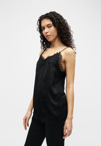 Γυναίκα με σγουρά μαλλιά που φοράει μαύρο τοπάκι με λεπτές τιράντες και δαντελωτή διακόσμηση, καθώς και μαύρο παντελόνι, στέκεται μπροστά σε απλή ανοιχτόχρωμη φόντο.