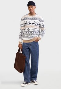 Maglione lavorato a maglia color crema con motivi geometrici blu navy e beige, abbinato a jeans di denim blu e sneakers nere, con una borsa a tracolla marrone in mano.