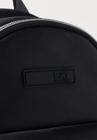 Svart nylonryggsäck med en slät yta, silverdragkedjebeslag och en matt svart logotypapplikation präglad med "EA7 Emporio Armani."