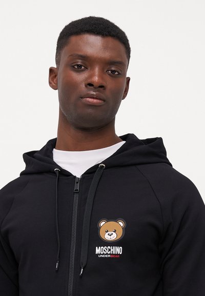 Black zip-up hoodie featuring a bear graphic on the chest, with "Moschino Under Bear" text. Soft fabric and adjustable drawstring hood.