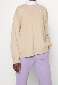 Sweat-shirt beige en tissu doux, avec un petit logo sur la poitrine, manches longues et coupe décontractée, associé à un pantalon lavande.
