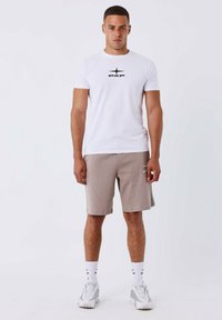 Witte korte mouwen t-shirt met een zwart logo, gecombineerd met beige shorts. Het model draagt ook witte sokken en grijze sneakers. Eenvoudig design.