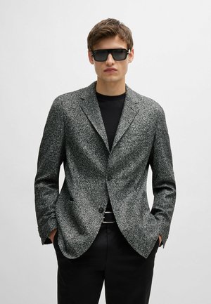Blazer grigio a spina di pesce con texture, revers a punta, chiusura con un solo bottone e due tasche frontali, indossato sopra una camicia nera.