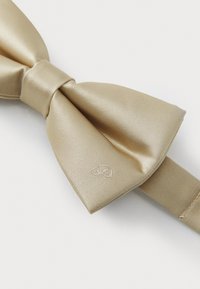 Nœud papillon en satin doré avec une finition texturée, présentant un petit détail de logo sur le côté et une sangle ajustable pour un ajustement sécurisé.