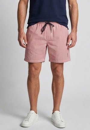 Man wearing red and white checkered shorts with black drawstring, navy shirt, and white sneakers, standing against a gray background.