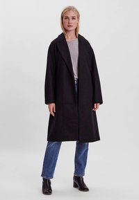 Cappotto lungo nero con collo a revers, realizzato in tessuto strutturato. Indossato sopra una maglia grigio chiaro e jeans blu, con tasche laterali.
