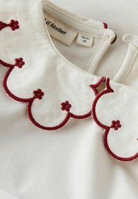 Collar de tela blanca con borde ondulado, con bordado floral rojo. Incluye un detalle de cremallera decorativa en la parte posterior.