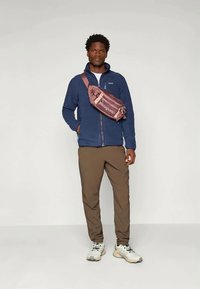 Uomo con giacca di pile blu navy, pantaloni marroni, sneakers bianche e una borsa a tracolla rosa Patagonia, in piedi davanti a uno sfondo neutro.