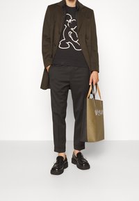 Cappotto verde oliva, maglione nero con grafica, pantaloni neri, mocassini neri e una borsa a spalla verde oliva con manici a contrasto e dettagli con logo.
