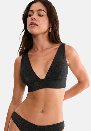 Femme portant un bikini noir scintillant avec un haut à col en V profond et un bas assorti, posant avec un bras dans le dos et portant des boucles d'oreilles créoles dorées.
