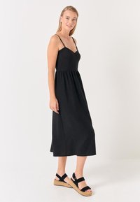 Abito maxi nero con spalline sottili, busto aderente e gonna fluida. Abbinato a sandali neri con cinturini su uno sfondo neutro.
