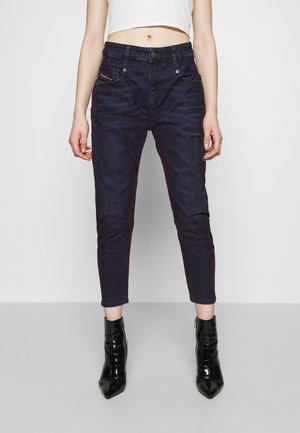 Frau trägt dunkelblaue, hoch taillierte, verkürzte Jeans und schwarze Lackleder-Stiefeletten, mit einem weißen, kurzen Oberteil.