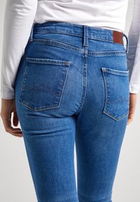 Jeans ajustados de mezclilla azul con cintura alta. Presentan dos bolsillos traseros con un sutil detalle de costura puntiaguda y un parche de cuero marrón en la cinturilla.