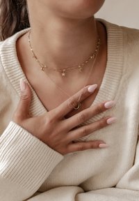 Collares dorados en capas con charms de estrella y luna sobre un suéter texturizado; uñas de color rosa pálido visibles en una mano que descansa sobre el pecho.