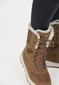 Bruna isolerade kängor med vit syntetisk päls vid kragen, bruna snören och texturerade paneler. Har en dämpad sula och sömnadsdetaljer.