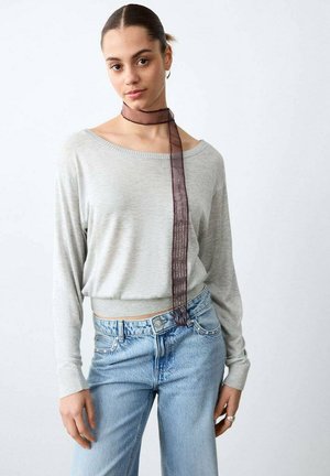 Giovane donna con indosso una maglia a maniche lunghe grigio chiaro, jeans blu, una sciarpa leggera color viola scuro annodata al collo e un anello d'argento sulla mano destra.