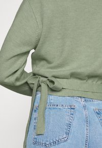 Zielony sweter dziany, związany w kokardę w talii, na jasnoniebieskich dżinsach z widocznymi szwami na tylnych kieszeniach.