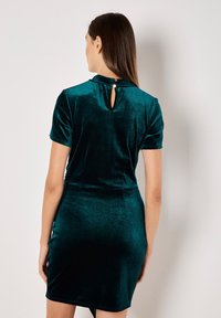 Samt Kleid in Dunkelteal mit kurzen Ärmeln, tailliertem Schnitt, Ausschnitt am Rücken und leichtem Schwung am Saum. Glatte Textur, elegantes Erscheinungsbild.