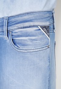 Jeans de denim azul claro con un bolsillo definido, costuras sutiles y una etiqueta azul oscuro con texto blanco. Textura suave y diseño ajustado.