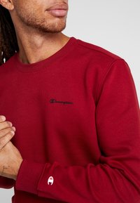 Felpa rossa con colletto girocollo, materiale leggero e logo "Champion" nero. Presenta un accento bianco del logo sulla manica.