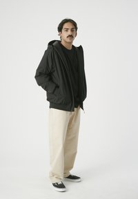 Schwarz-hooded Jacke mit Reißverschluss, getragen über einem schwarzen Hemd, kombiniert mit beigen, weiten Hosen und schwarzen Sneakers mit weißen Akzenten.