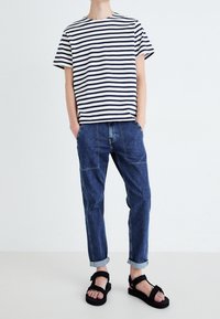 Camiseta a rayas en azul marino y blanco, combinada con pantalones denim oscuros enrollados y sandalias negras de tiras. Estilo casual, ajuste relajado.