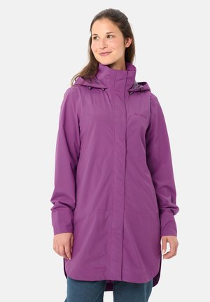 Femme portant un long imperméable violet à capuche, debout devant un fond gris clair uni, regardant sur le côté avec un léger sourire.