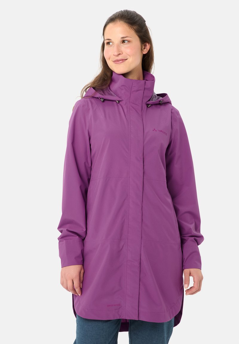 Femme portant un long imperméable violet à capuche, debout devant un fond gris clair uni, regardant sur le côté avec un léger sourire.