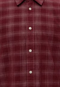 Chemise en velours côtelé marron rouge avec motif à carreaux blanc et rose clair et boutons blancs au centre devant sous un col pointu.