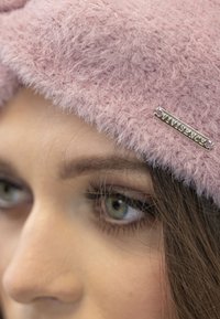 Gorro rosa esponjoso con superficie texturada, que presenta una etiqueta metálica plateada con la inscripción "VIVISENCE". Enfoque en la mitad superior de la cara.