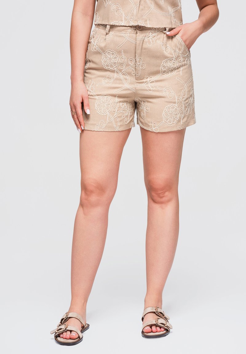 Short beige avec broderie florale blanche porté avec un haut assorti et des sandales beiges, une main dans la poche et l'autre reposant sur le côté.
