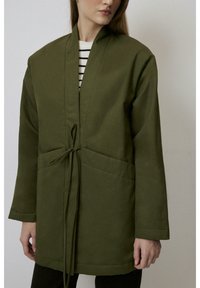Jacheta de stil kimono, de culoare verde măslin, cu mâneci lungi, închidere prin legare și două buzunare frontale, realizată dintr-un material neted cu un finisaj mat.