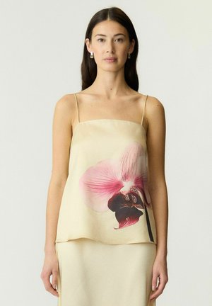 Femme aux longs cheveux foncés porte un haut sans manches crème et une jupe avec un grand motif orchidée rose et rouge foncé sur le haut.