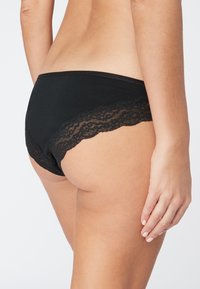 Culottes noires avec dentelle, finition lisse, présentant des bords festonnés et un tissu doux. La vue latérale souligne le design élégant.