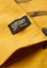 Tessuto giallo senape con una superficie texture, caratterizzato da un'etichetta visibile blu navy "SUPERDRY MFG CO." e motivi neri a contrasto.
