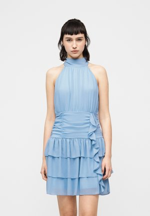 Mladá žena v bezrukávových svetlomodrých šatách s volánmi na vrstvených riaseniach a detailom na zhromaždení v páse, stojaca pred jednoduchým pozadím.