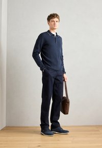 Marine langermet polo med krage, marinebukser og mørke sko. Brun budveske med skulderrem. Tregulv og nøytral bakgrunn.