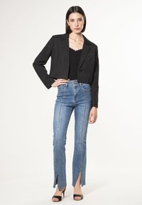 Giacca nera sfilettata con risvolti, indossata sopra un top nero. Jeans a vita alta blu chiaro con fessure laterali e cuciture a contrasto. Tacchi neri.