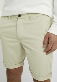 Shorts en coton beige avec ourlets retroussés, poches avant et fermeture à bouton. Texture lisse avec des détails de couture subtils.