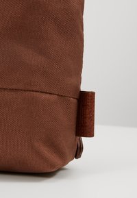 Coin d'un sac en tissu marron avec une petite boucle en cuir attachée sur le côté sur un fond clair uni.