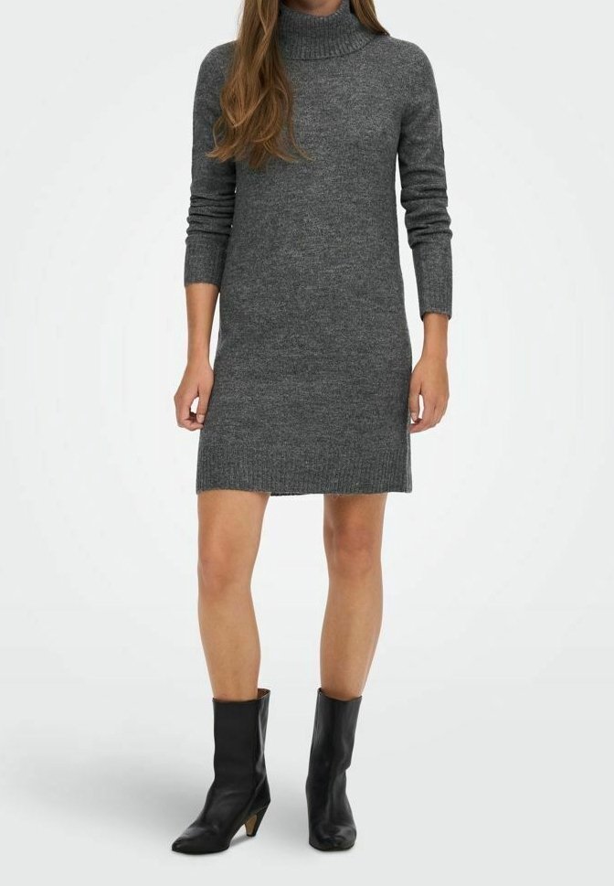 Femme portant une robe en maille grise à col roulé et manches longues, et des bottes noires à talons mi-mollet, debout devant un fond blanc uni.