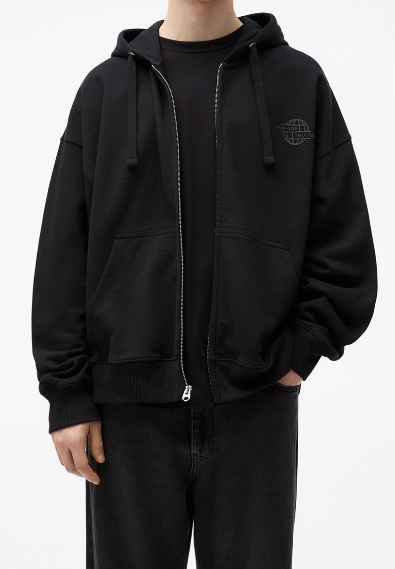 Homme portant un sweat à capuche noir zippé avec poches avant, cordons de serrage et un petit logo globe sur la poitrine, associé à un pantalon noir.